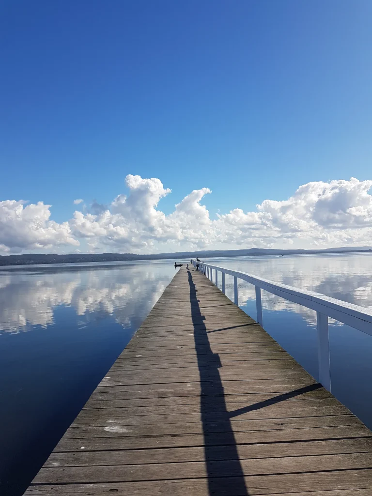 Long Jetty stretches across the calm waters of Tuggerah Lake in the Central Coast, reflecting fluffy white clouds beneath a bright blue sky. This peaceful waterfront spot is one of the best day trips from Sydney, offering scenic lake views, leisurely walks, and a relaxing escape just north of the city.