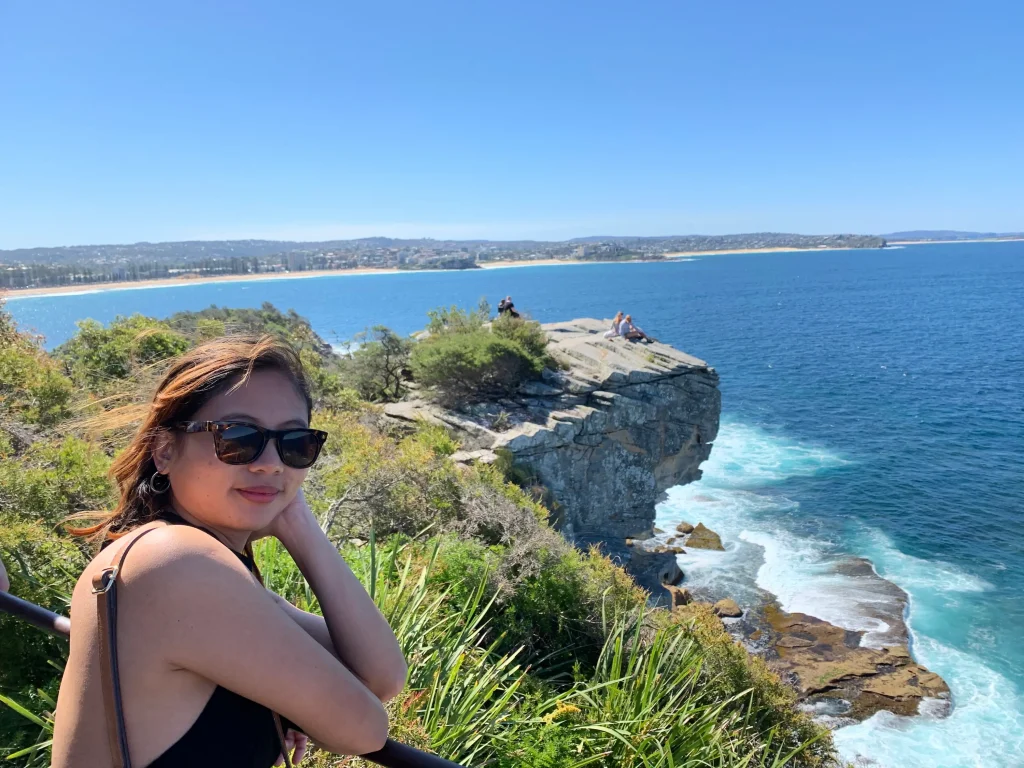 A traveler overlooks the rugged sandstone cliffs of Manly as turquoise waves crash against the rocky shoreline under a clear blue sky. This scenic coastal lookout is one of the best places to visit in Sydney, with spectacular ocean views and walking trails along the city's northern beaches.