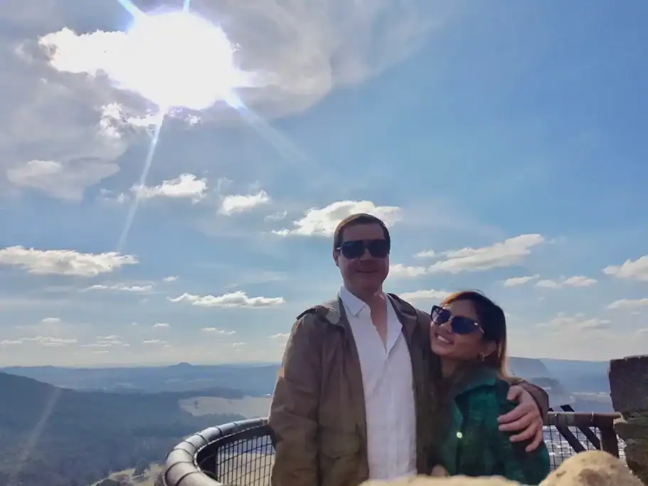 A couple stands at Mount Jellore Lookout in Bowral, overlooking rolling hills, lush bushland, and distant valleys beneath dramatic clouds with sunlight breaking through the sky. This peaceful lookout is one of the most scenic places to visit outside Sydney, offering panoramic views of the Southern Highlands on an easy day trip.