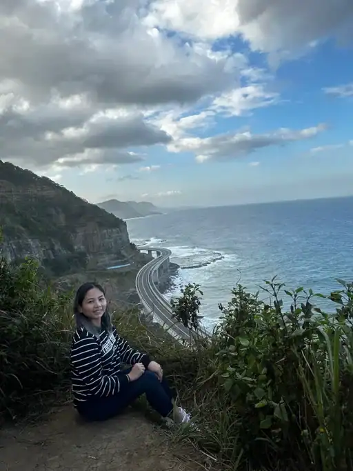 A traveler sits at a cliffside lookout above the Sea Cliff Bridge with dramatic ocean views, towering coastal cliffs, and thick clouds stretching across the sky. This scenic stop along the Grand Pacific Drive is one of the most iconic things to do outside Sydney, combining spectacular coastal scenery with one of Australia's most picturesque ocean roads.