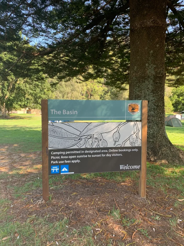 Welcome sign for The Basin picnic and camping area in Ku-ring-gai Chase National Park surrounded by grassy grounds and tall Norfolk pine trees. The sign reads, "The Basin. Camping permitted in designated area. Online bookings only. Picnic Area open sunrise to sunset for day visitors. Park use fees apply. Welcome." The Basin is one of the most relaxing places to visit outside Sydney for picnics, camping, and waterfront walks.