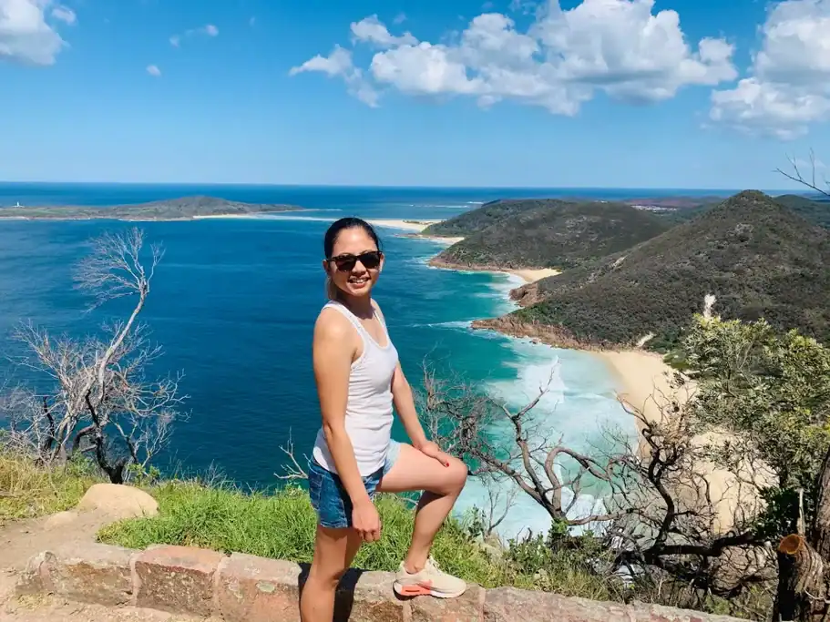 A traveler poses at Tomaree Head Lookout in Port Stephens with panoramic views of turquoise water, golden beaches, and rugged coastal headlands under a bright blue sky. This iconic lookout is one of the best day trips from Sydney, offering spectacular views and one of the most memorable coastal walks in New South Wales.