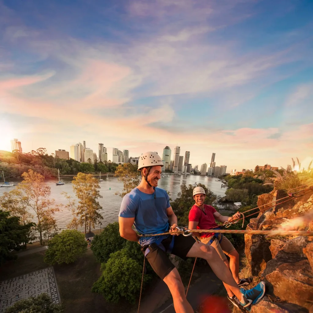 Two climbers abseiling down the Kangaroo Point Cliffs at sunset with the Brisbane River and skyline behind them. For travellers wondering what to do in Brisbane, abseiling offers an adventurous way to experience the city from above.