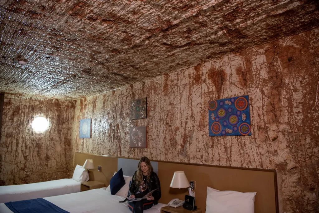 Woman sitting on a bed inside a room carved into the red rock at the Desert Cave Hotel. Staying in an underground hotel is one of the most unique things to do in Coober Pedy.