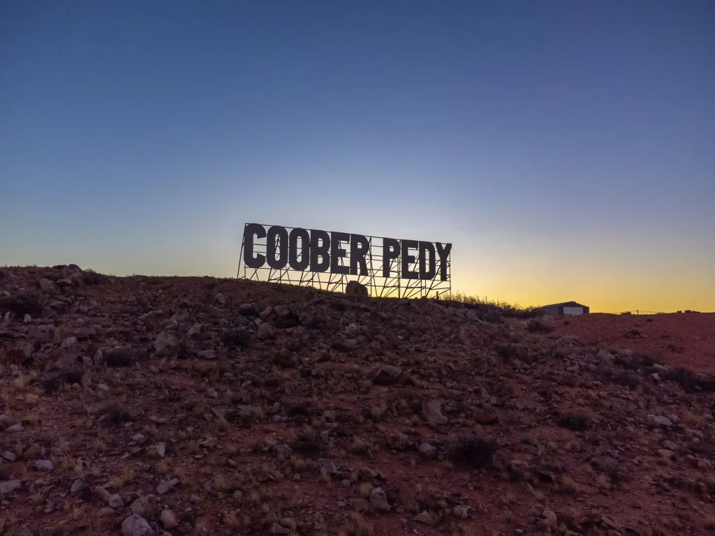 Large "COOBER PEDY" sign silhouetted against a golden sunset above the rocky desert landscape. Seeing the outback town at sunset is one of the memorable things to do in Coober Pedy.