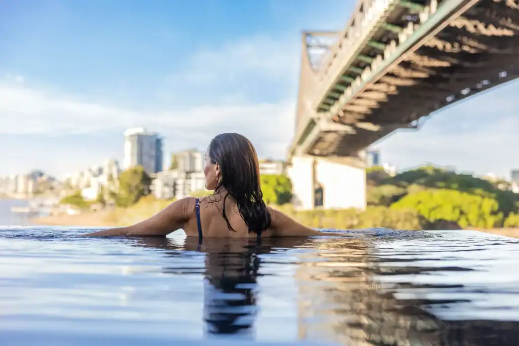 Woman relaxing in the Crystalbrook Vincent hotel infinity pool with the Story Bridge stretching overhead and Brisbane city views in the distance. The riverside pool is a standout feature for travelers looking for a Brisbane hotel with a pool.