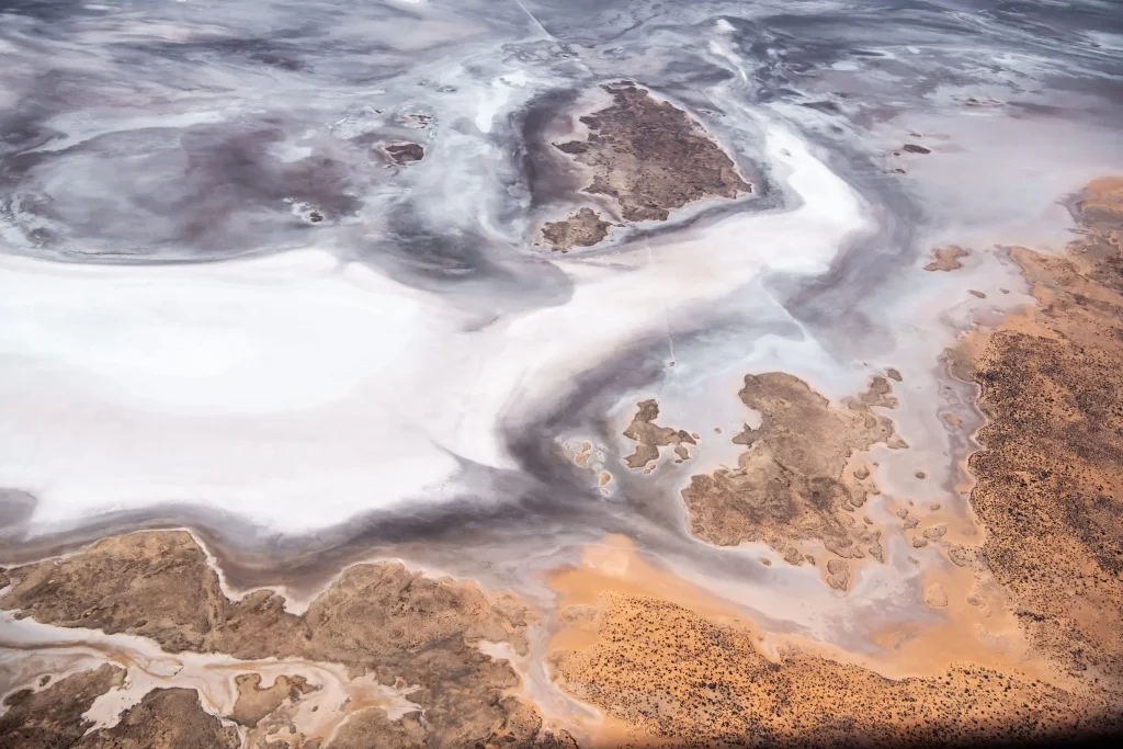 Aerial view of Lake Eyre with sweeping white salt flats and swirling grey patterns surrounded by orange brown desert. A scenic flight over Lake Eyre is a memorable activity to add to a Coober Pedy itinerary.
