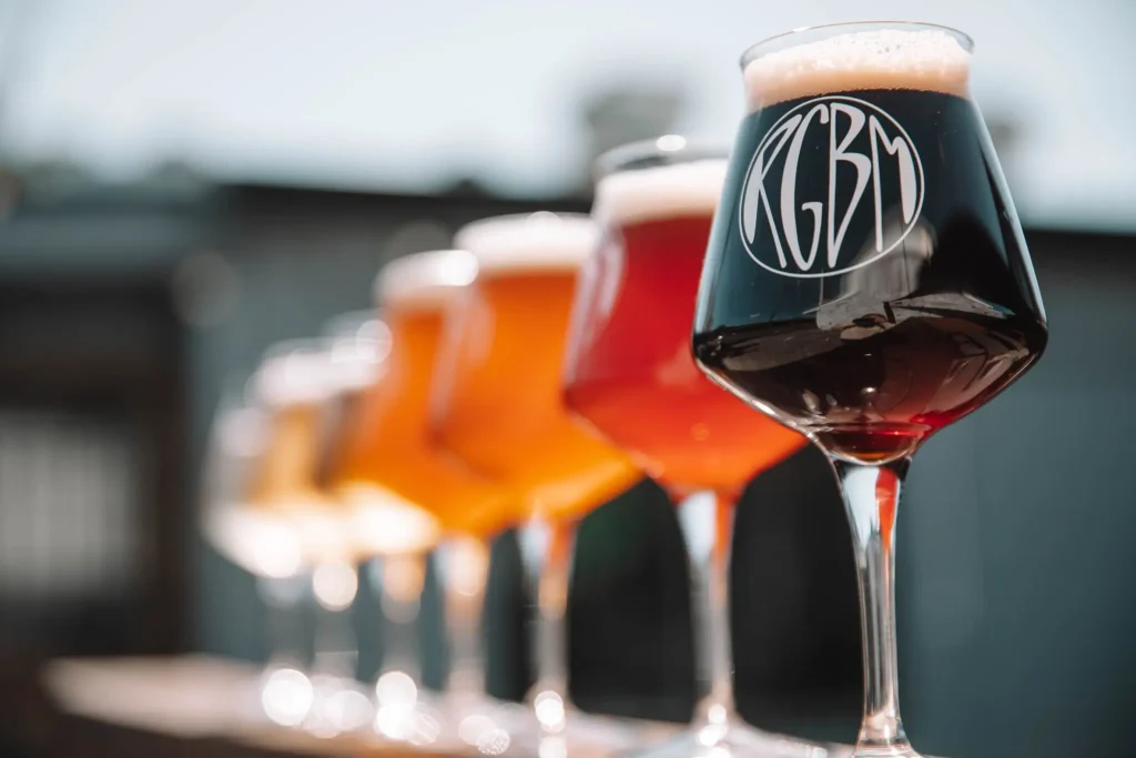A row of tulip glasses filled with craft beers ranging from pale gold to deep black is lined up for a tasting at Reub Goldberg Brewing Machine. The NSW craft beer scene is showcased through a variety of beer styles served for visitors to sample.