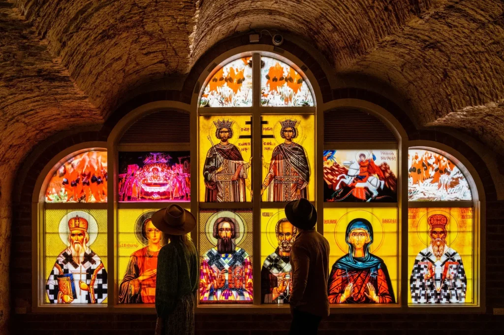 Two visitors in wide brimmed hats admire glowing stained glass religious icons inside the underground Serbian Orthodox Church. Visiting this rock carved church is one of the most distinctive Coober Pedy attractions.