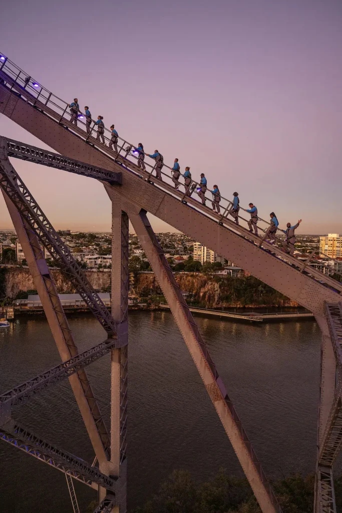 Climbers in safety harnesses ascending the steel arches of Brisbane Story Bridge at dusk with the river and city below. The Story Bridge Adventure Climb is a memorable addition to a Brisbane itinerary for panoramic views from above the city.