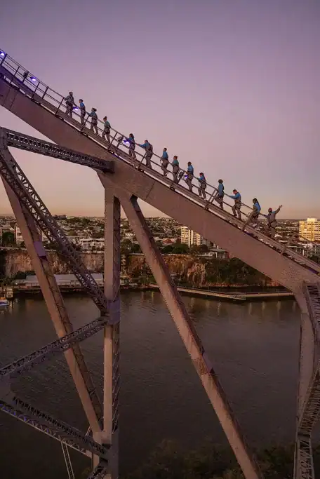 Climbers in safety harnesses ascending the steel arches of Brisbane Story Bridge at dusk with the river and city below. The Story Bridge Adventure Climb is a memorable addition to a Brisbane itinerary for panoramic views from above the city.
