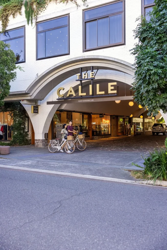 The arched entrance of The Calile Hotel in Brisbane with its name displayed in large gold letters above the driveway. Two people stand with bicycles outside the leafy hotel which is featured among the best hotels in Brisbane with a pool.