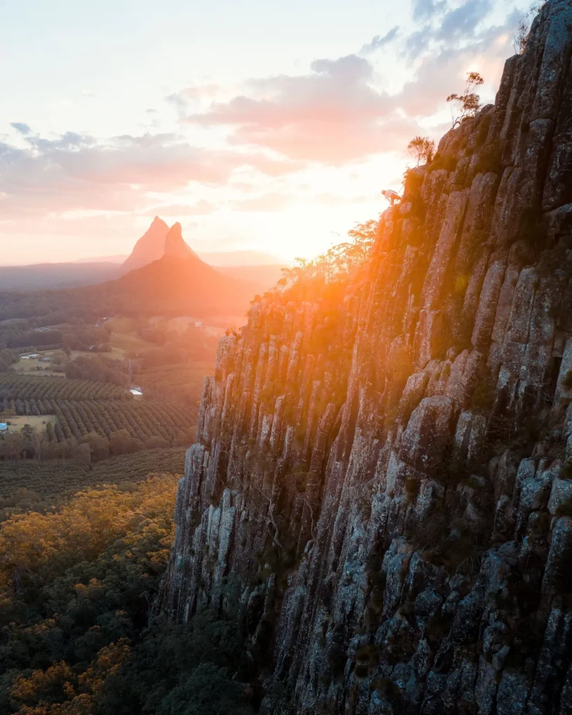 Steep rocky cliffs glowing in the sunset with the distinctive peaks of the Glass House Mountains rising over the forested landscape. Hiking in the Glass House Mountains is one of the adventurous things to do near Brisbane for dramatic views.