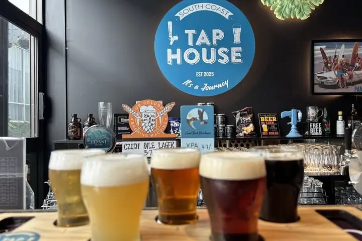 A tasting paddle of craft beers sits on the bar at South Coast Tap House with taps and beer displays in the background. One of the South Coast breweries featured on the ale trail, the venue offers a range of locally brewed beers in a relaxed tasting setting.