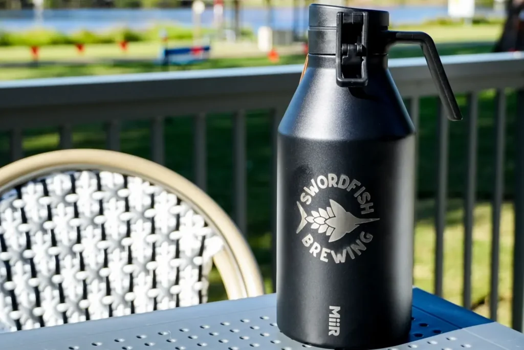 Black insulated growler with the Swordfish Brewing logo sits on an outdoor table overlooking the water. A refillable beer growler from a stop on the South Coast Ale Trail highlights the region's independent craft brewery scene.