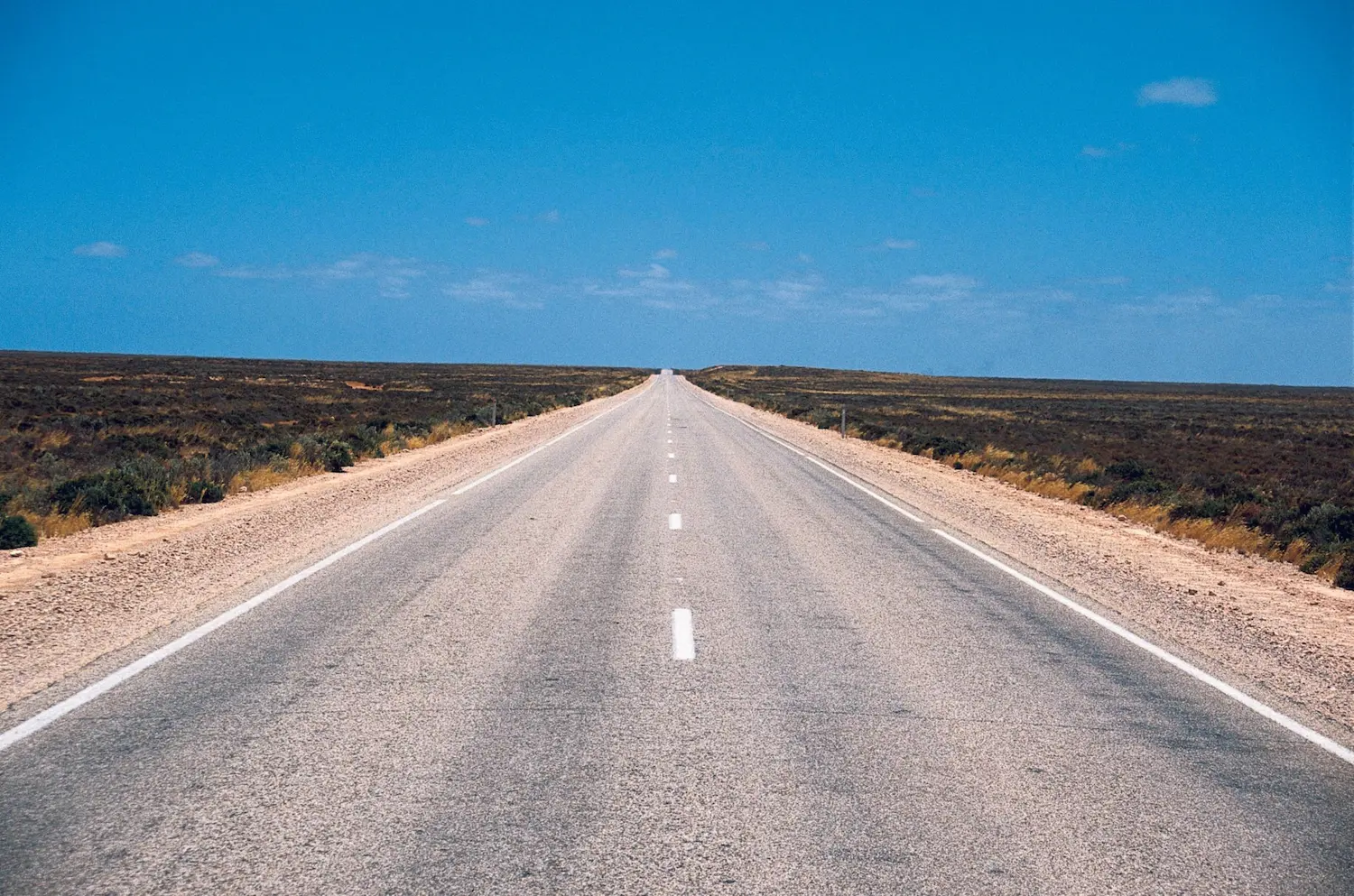 Long straight stretch of the Eyre Highway disappearing into the horizon beneath a clear blue sky. This remote highway forms part of the overland drive from Brisbane to Perth across Australia.