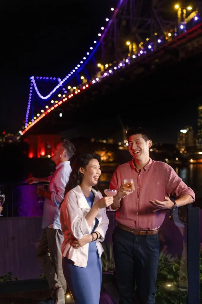 A couple laughs over cocktails at Fiume Bar at the Crystalbrook Vincent Hotel with the illuminated Story Bridge towering above them. The colourful nighttime setting captures the atmosphere of rooftop bars in Brisbane with river and landmark views.