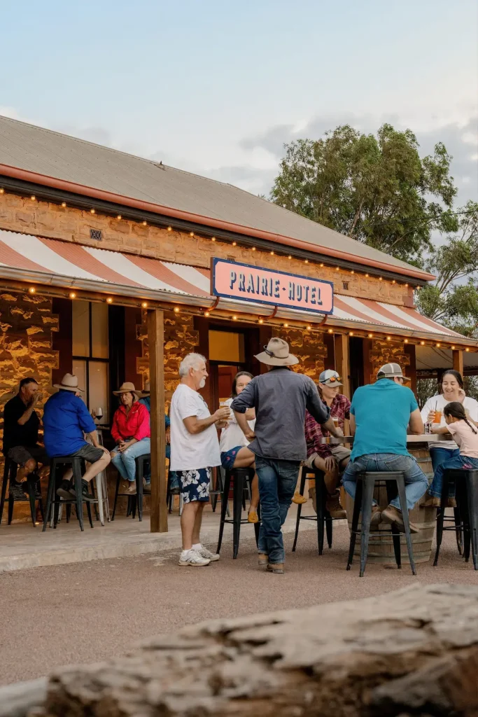 Travelers gather for drinks outside the historic stone Prairie Hotel beneath the sign reading "Prairie Hotel". A lively outback stop during a Flinders Ranges camping trip.