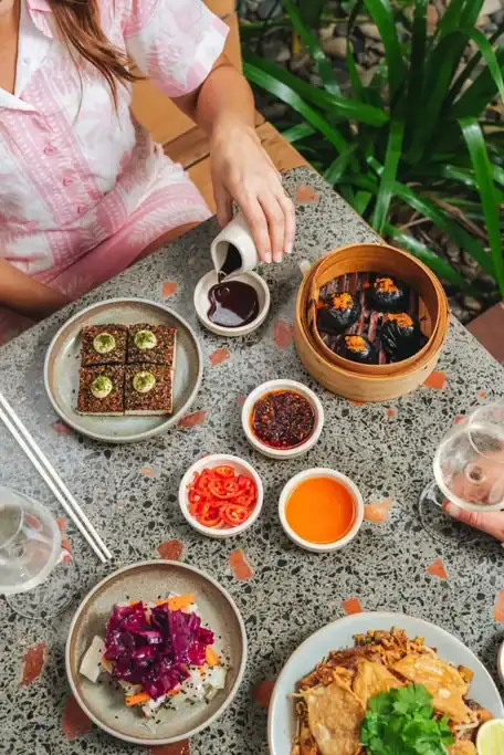 Shared meal at Southside Restaurant in Brisbane with black dumplings in a bamboo steamer. Colourful Asian inspired dishes and dipping sauces cover the table as diners pour sauce and enjoy wine.