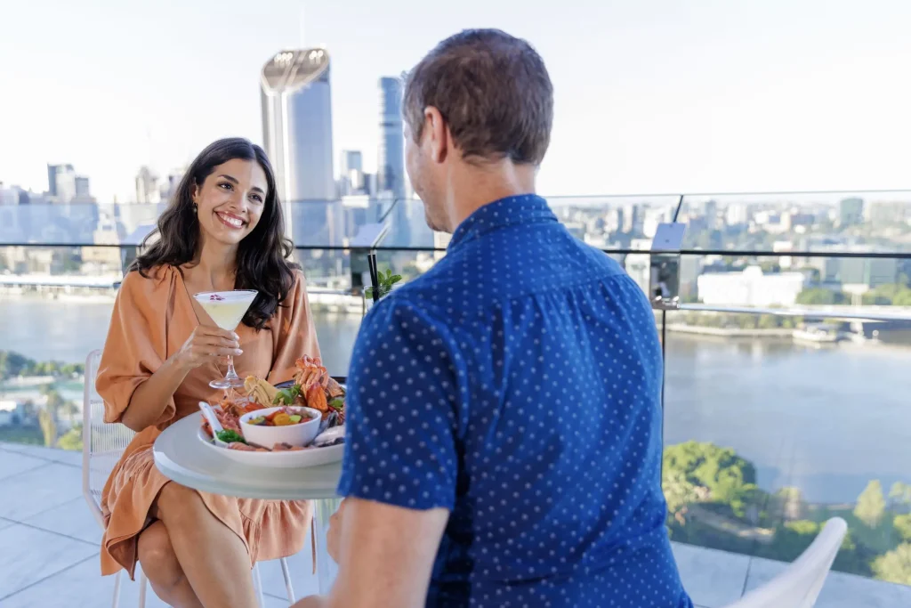 A couple enjoys cocktails and a seafood platter at the Emporium Hotel rooftop bar overlooking the Brisbane River and city skyline. The scenic terrace makes it a standout among the best rooftop bars in Brisbane.
