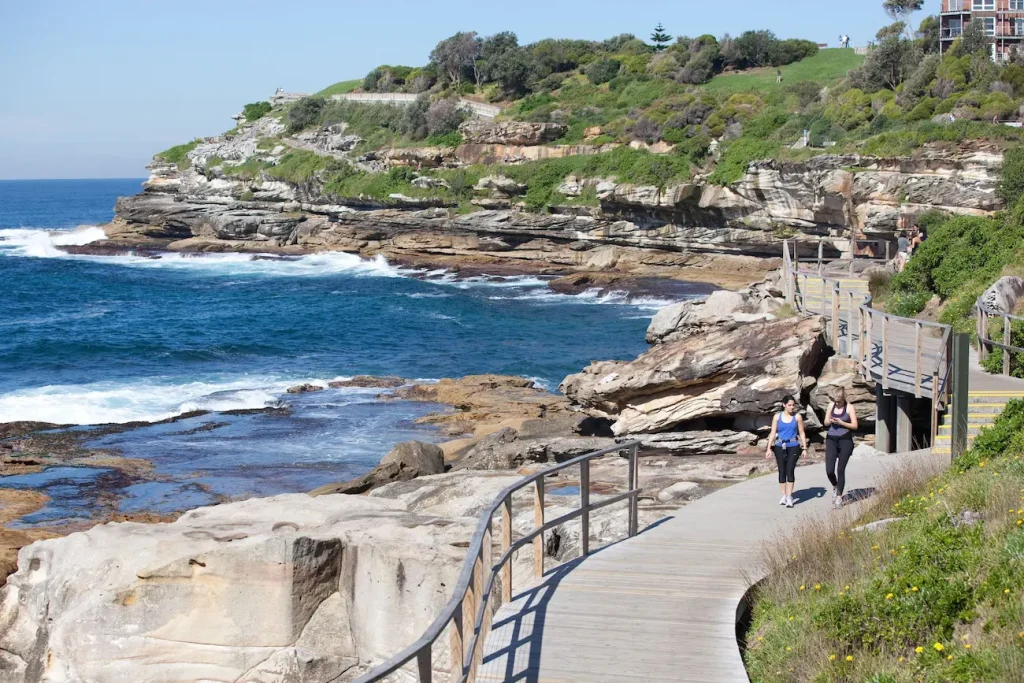 Two women walk along the Bondi coastal path beside sandstone cliffs and bright blue ocean. During whale migration season the walk is a free alternative to whale watching cruises in Sydney with opportunities to spot whales from shore.