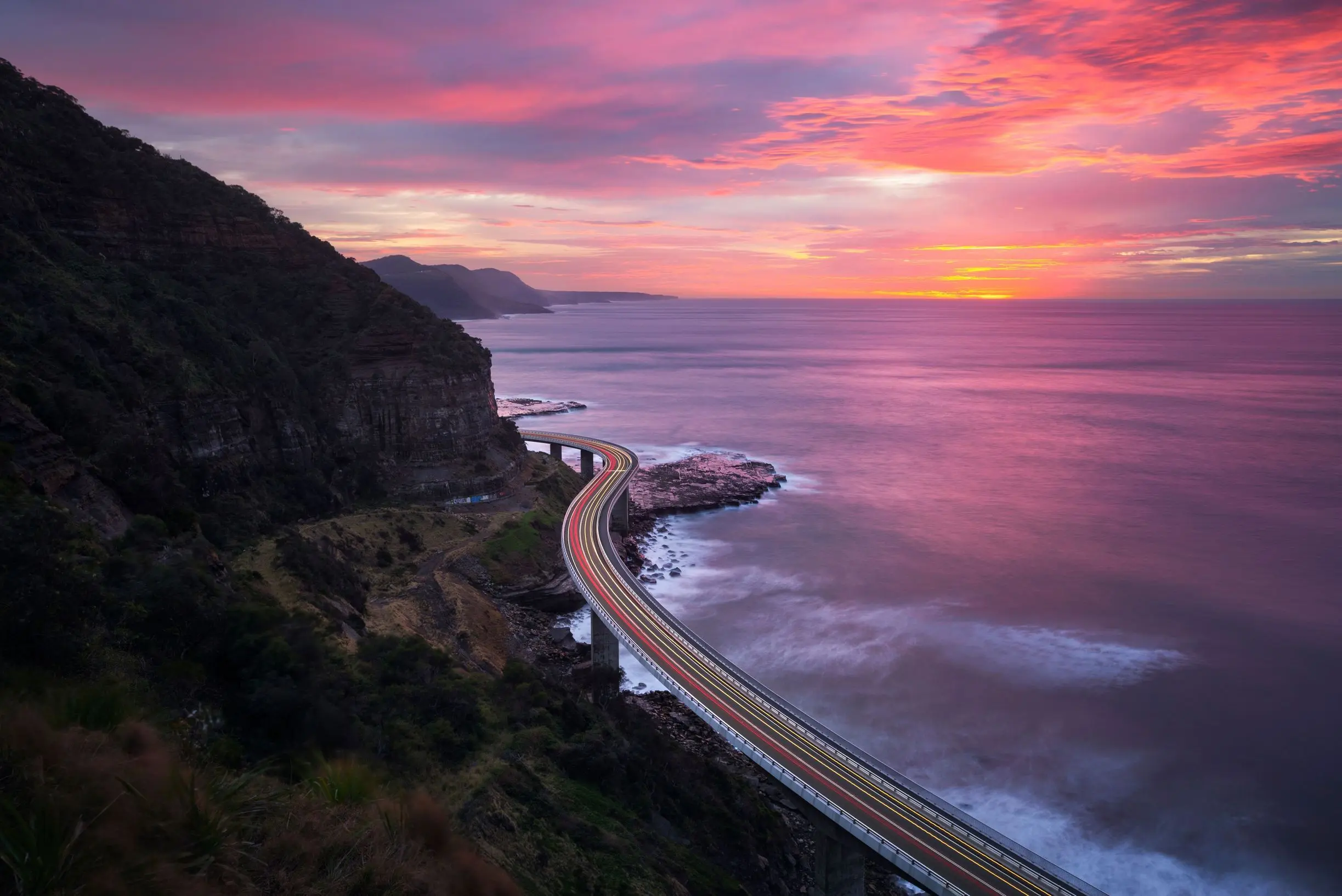 Sea Cliff Bridge curves between the steep coastal cliffs and ocean at Clifton beneath a vivid pink and orange sunset. Light trails along the bridge lead toward the coastline seen from the Sea Cliff Bridge