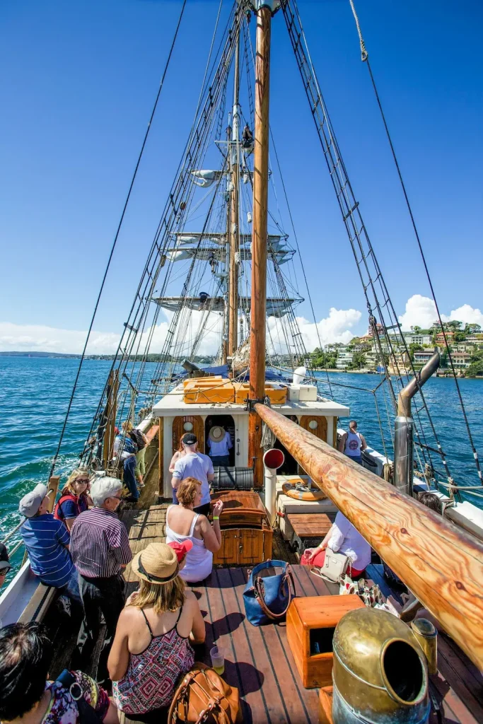 Passengers relax on the wooden deck of a tall ship sailing across the blue waters of Sydney Harbour. Tall ship cruises offer a unique alternative to traditional whale watching tours in Sydney.