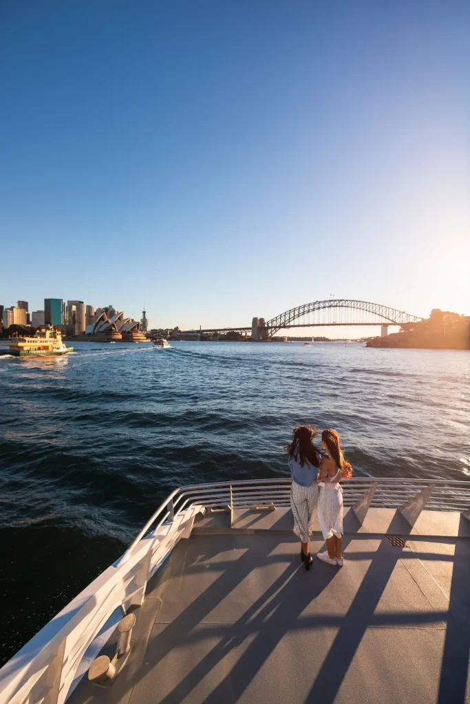 Two friends enjoy sunset views of the Sydney Opera House and Sydney Harbour Bridge on the return journey aboard a Captain Cook Cruises whale watching vessel. The harbour scenery is a memorable finish to a Sydney whale watching cruise.
